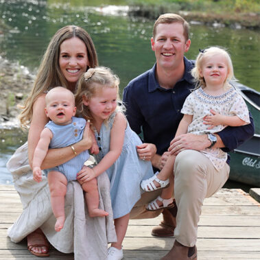 Smiling family of five posing on a wooden dock by a sunlit pond: mother with baby, two young daughters, and father with another daughter on his knee.