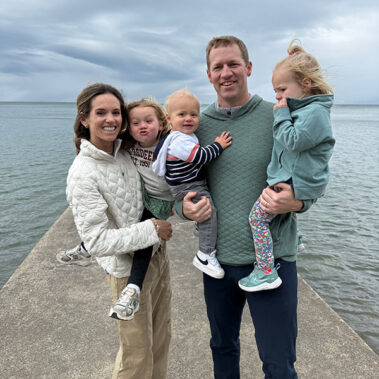 Family of five posing on a concrete pier by the water, smiling at the camera with a cloudy sky above.