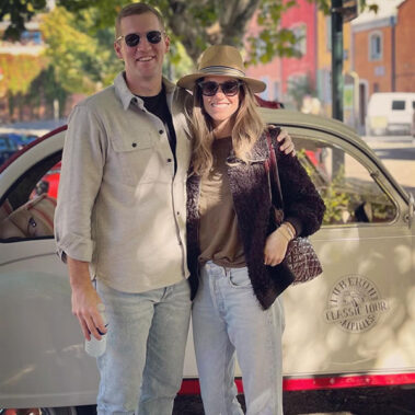 Couple smiling and posing together in front of a cream vintage car on a sunny street, with trees in the background.