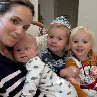 Mother on a sofa with her three children: baby in a whale-print onesie, a girl wearing a tiara, and a girl in an orange dress on the right.
