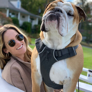 Smiling woman with sunglasses sits beside a large bulldog wearing a black harness on a sunny porch.