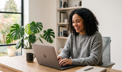 image 33-1 Woman with curly hair typing on a laptop at a wooden desk in a bright home office, with a plant and books nearby.